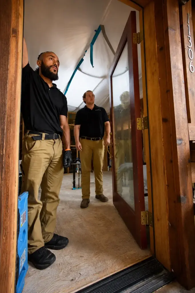 Two restoration technicians in black polo shirts and khaki pants inspecting the upper doorframe and ceiling of a home.