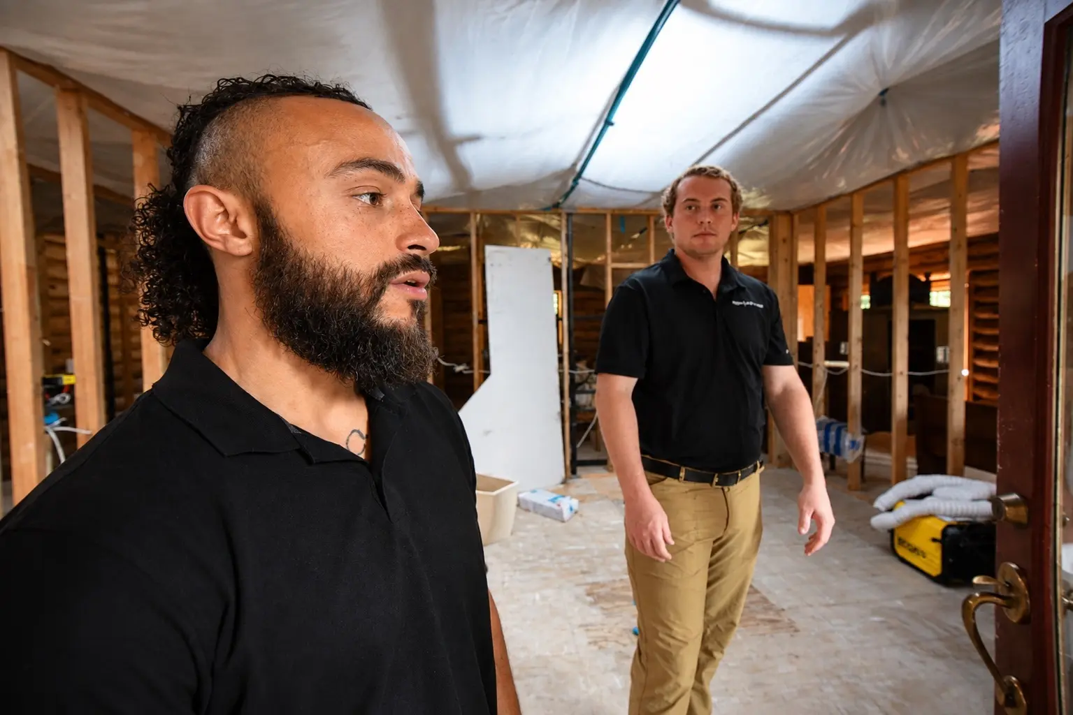 A bearded worker in profile in the foreground with a second worker in a black polo shirt and khaki pants standing behind him, inside a home restoration site featuring exposed wooden studs and plastic containment lining on the ceiling.